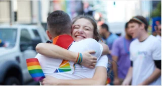 Employee experience: two people in white tshirts are doing a pride hug while holding a rainbow flag