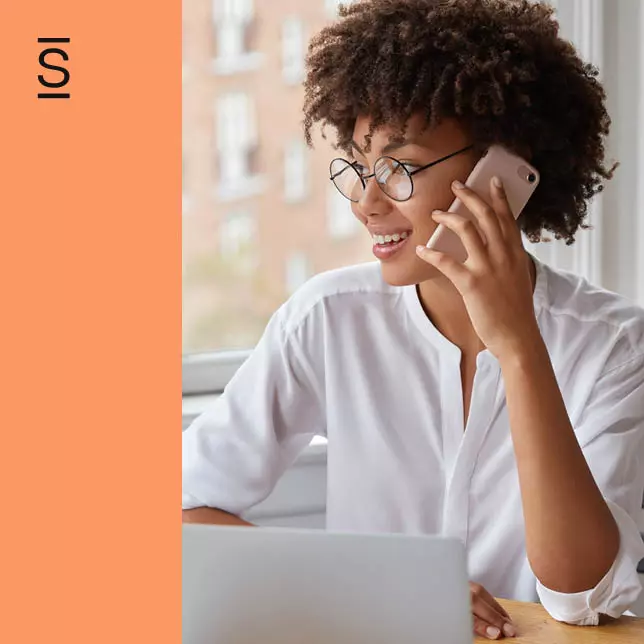 Manage Remote Teams - woman with glasses speaking on smartphone while using laptop computer and working in nonoffice location