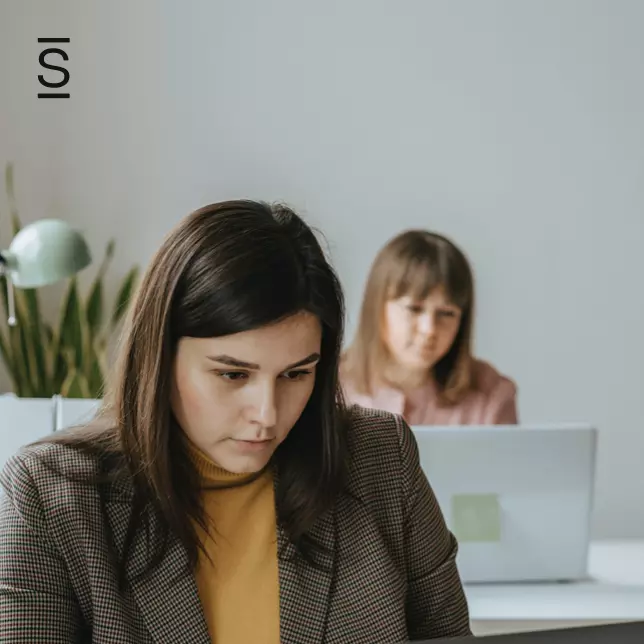 Intranet governance - woman in plaid blazer works on laptop while female colleague works behind her