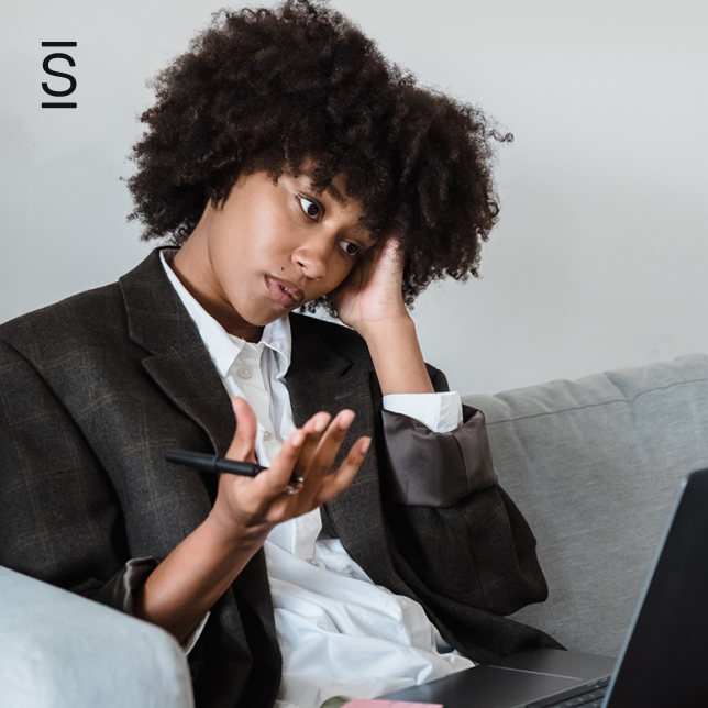 Employee experience - woman in blazer speaking on smartphone and looking at computer screen with confused look on her face