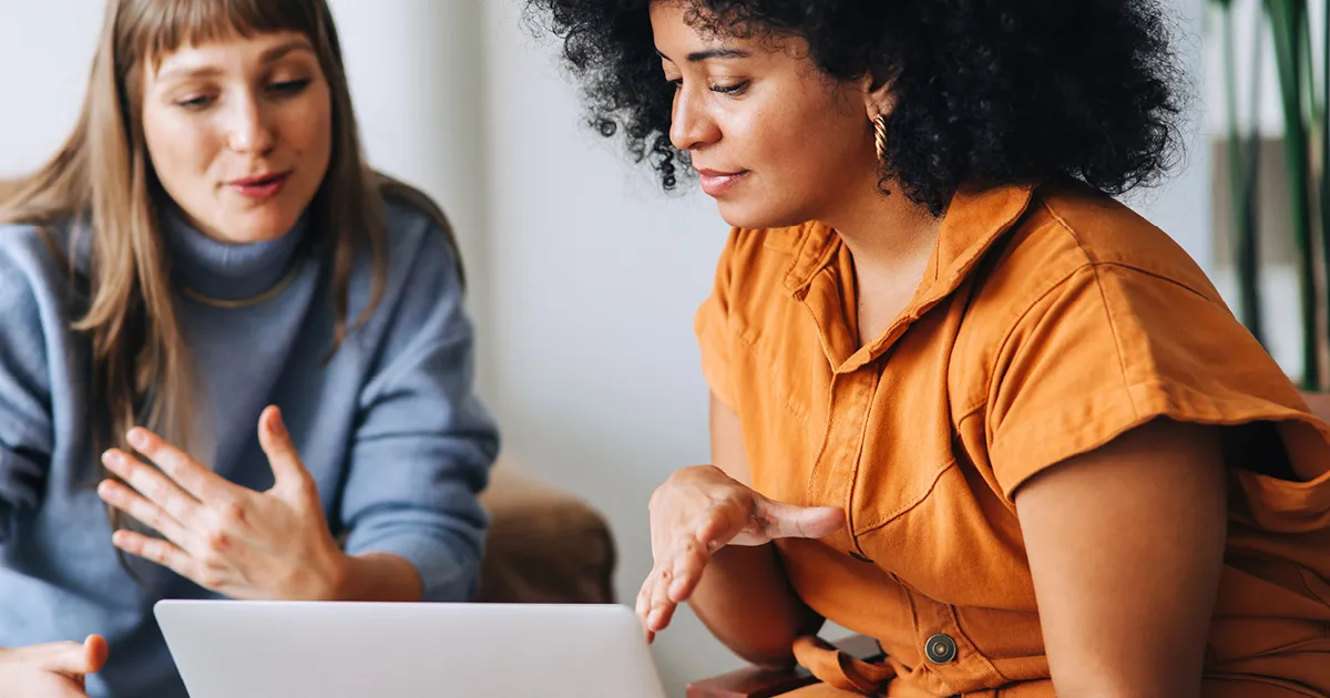 Employee retention - woman in grey shirt & woman in orange shirt speak while looking at laptop screen